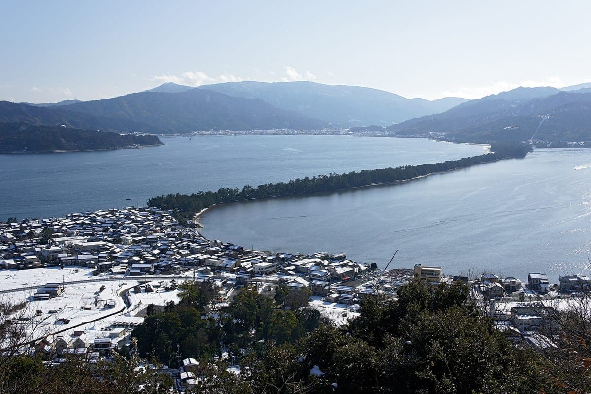 View from a hillside of the long pine-covered Amanohashidate sandbar stretching across a calm blue bay, with a town below and mountains beyond