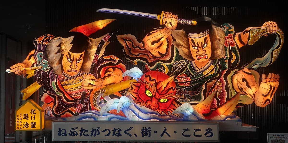 Una enorme carroza iluminada nebuta por la noche que muestra guerreros pintados con espadas, un demonio de cara roja y olas arremolinadas, brillando con colores brillantes contra un cielo oscuro.