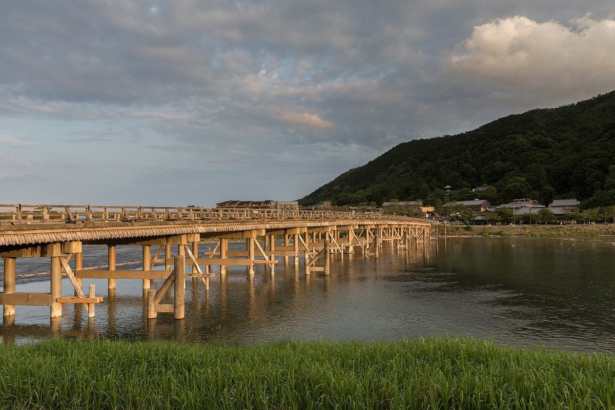 Le long pont en bois Togetsukyō enjambant la large rivière d'Arashiyama, avec derrière lui des collines boisées éclairées par le soleil de fin d'après-midi