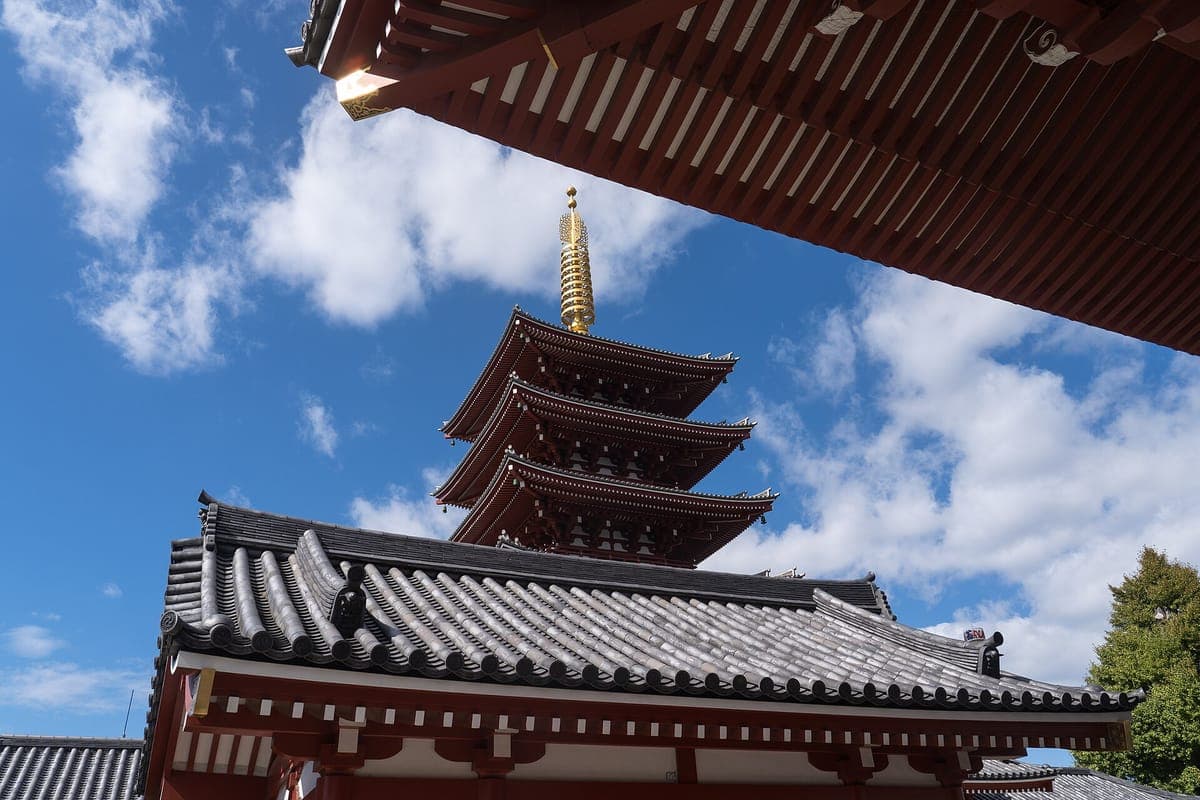 O pagode de cinco andares de Sensō-ji com uma torre dourada erguendo-se acima do telhado de um templo contra um céu azul com nuvens brancas