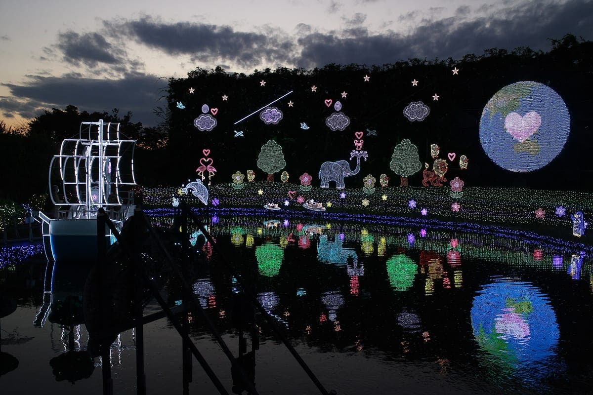 La iluminación invernal del Ashikaga Flower Park al anochecer: escenas LED de árboles, animales y un globo resplandeciente con un corazón reflejado en un estanque, junto a una escultura luminosa con forma de barco