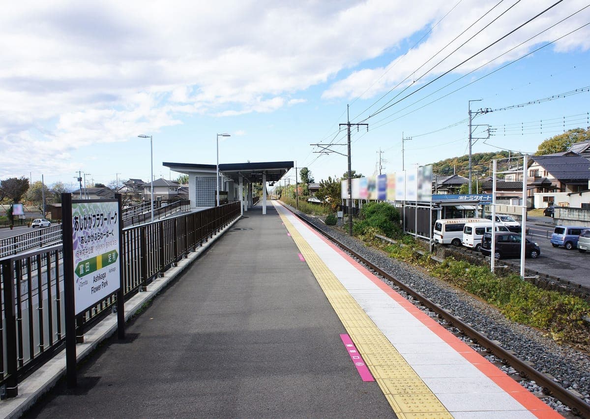 El andén único de la estación Ashikaga Flower Park de la línea JR Ryōmō, con una franja táctil amarilla, un cartel con el nombre de la estación y un pequeño refugio bajo un cielo azul.
