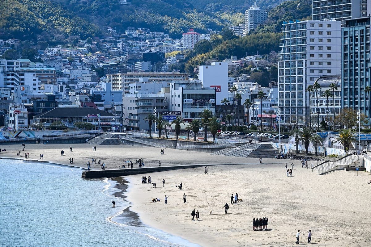 Atami Sun Beach, zakřivená písečná pláž lemovaná palmami, s hotely a bytovými domy tyčícími se do zelených kopců za