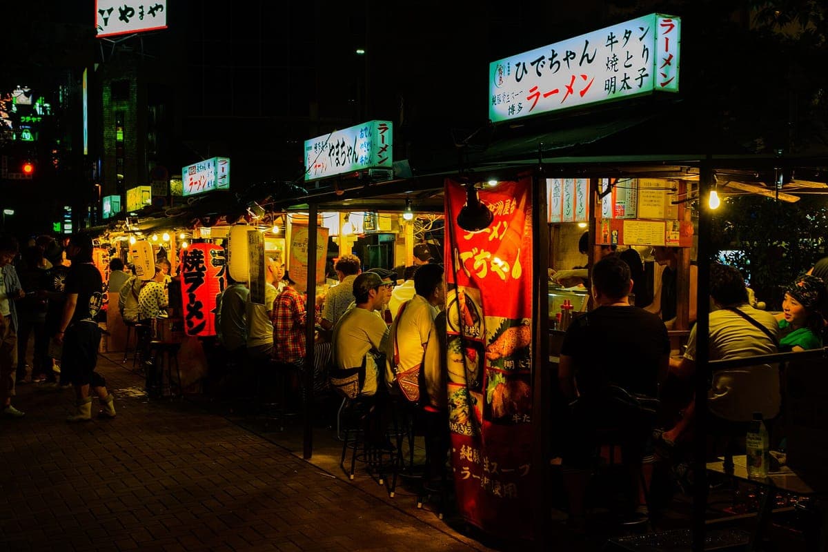 แผงขายอาหารเรียงรายสว่างไสวในเวลากลางคืนใน Nakasu ฟุกุโอกะ พร้อมม่านสีแดง ป้ายราเม็งเรืองแสง และผู้คนที่นั่งอยู่ที่เคาน์เตอร์