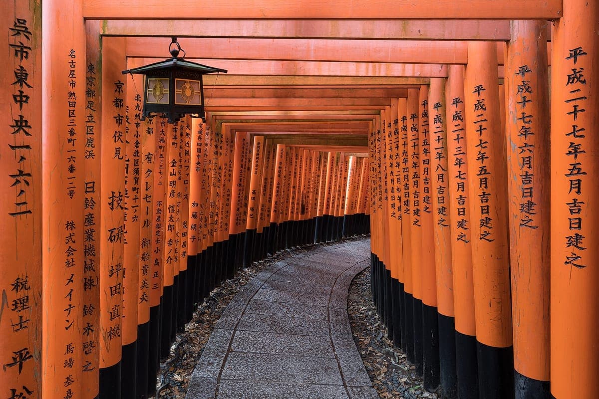 A path curving through a tunnel of vermilion torii gates at Fushimi Inari Taisha, with black-painted bases and a hanging lantern