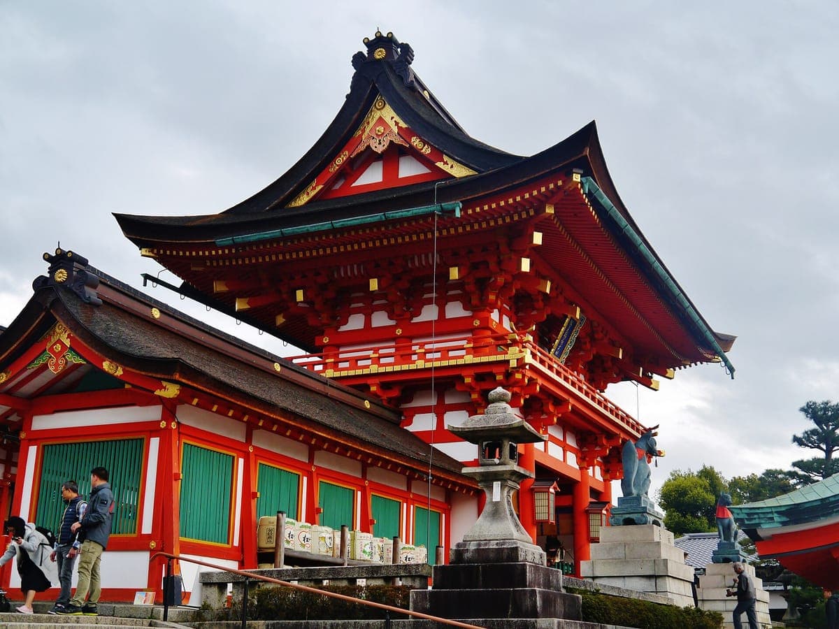 La puerta bermellón de dos pisos Rōmon de Fushimi Inari Taisha con paredes de celosía verde y una linterna de piedra al frente