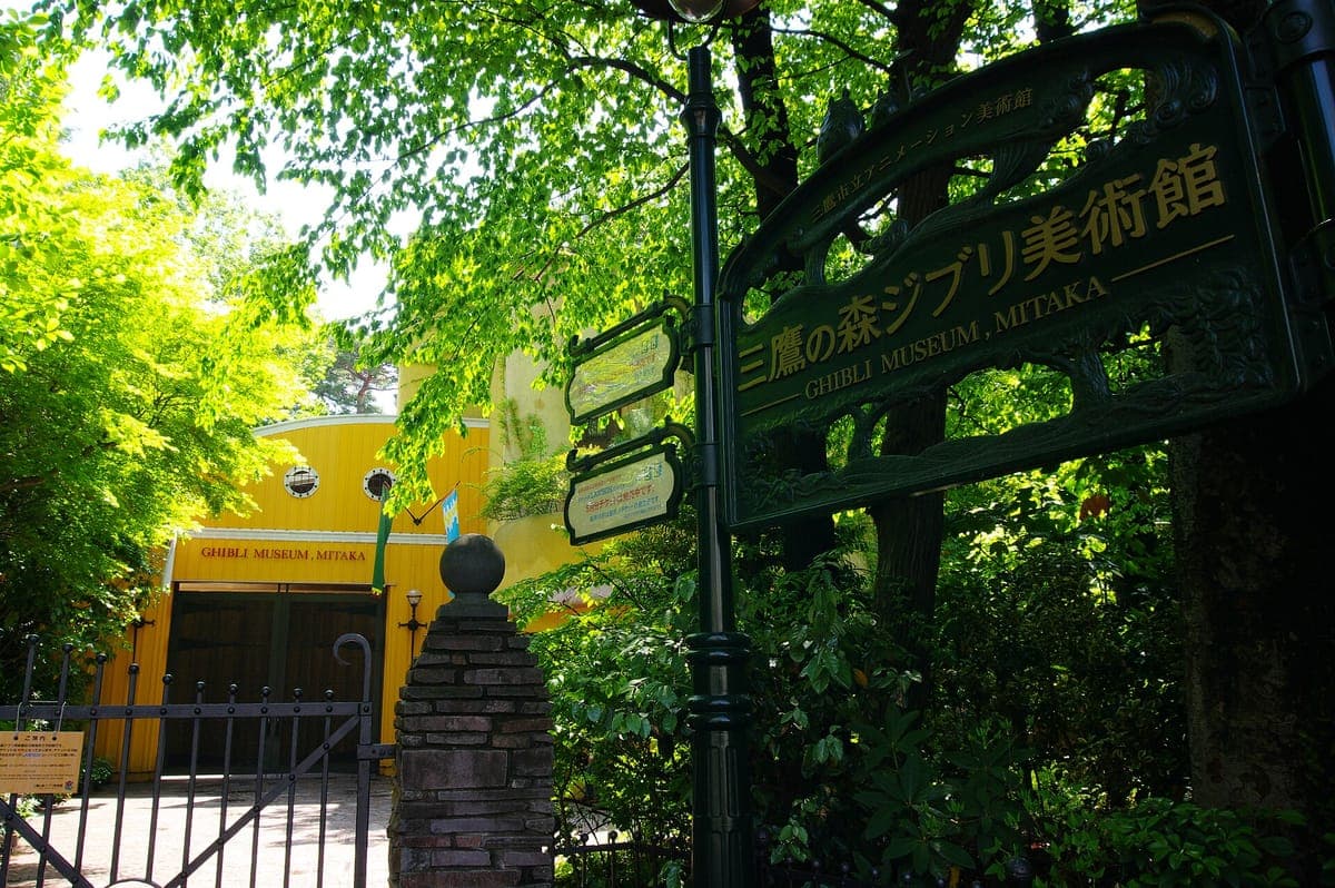 El cartel verde del Museo Ghibli, Mitaka, con su nombre japonés en letras doradas, bajo árboles verdes y frescos, con el edificio amarillo del museo detrás