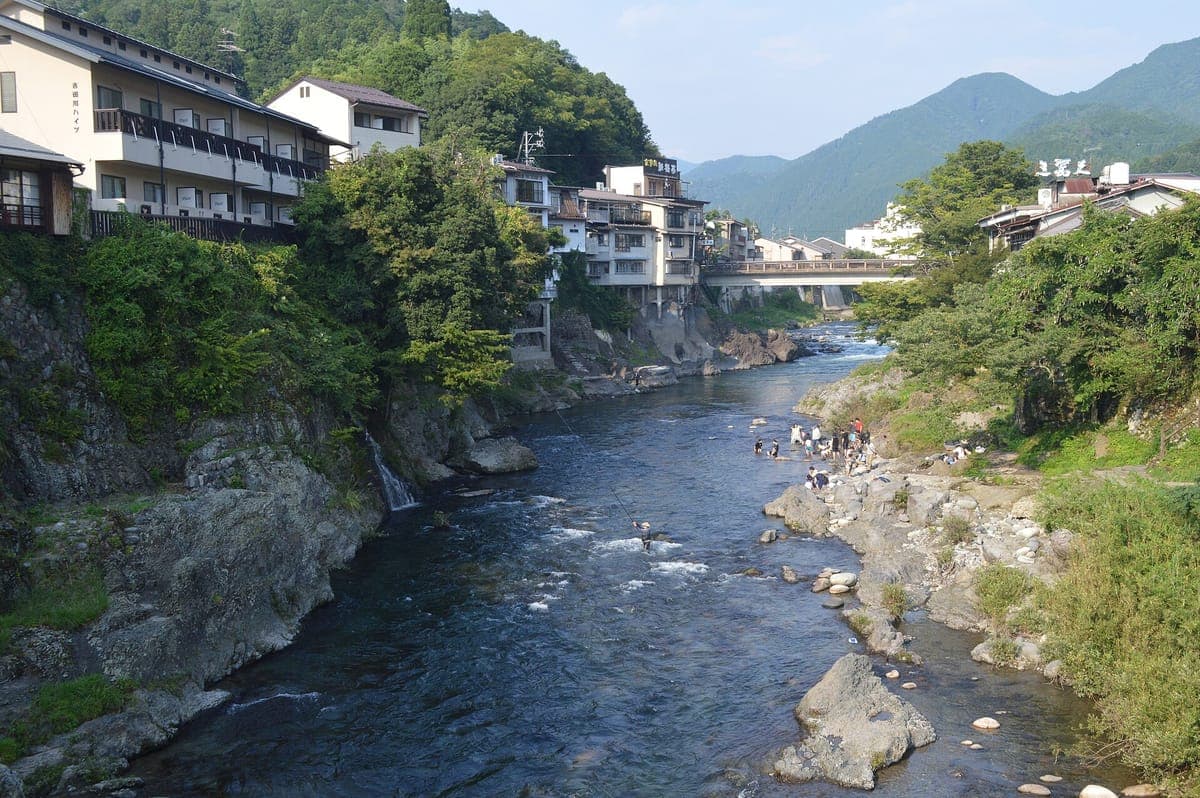 La rivière Yoshida, limpide, coule entre des rives rocheuses à Gujō Hachiman, avec des maisons construites au-dessus de l'eau et un pont et des montagnes verdoyantes au-delà.