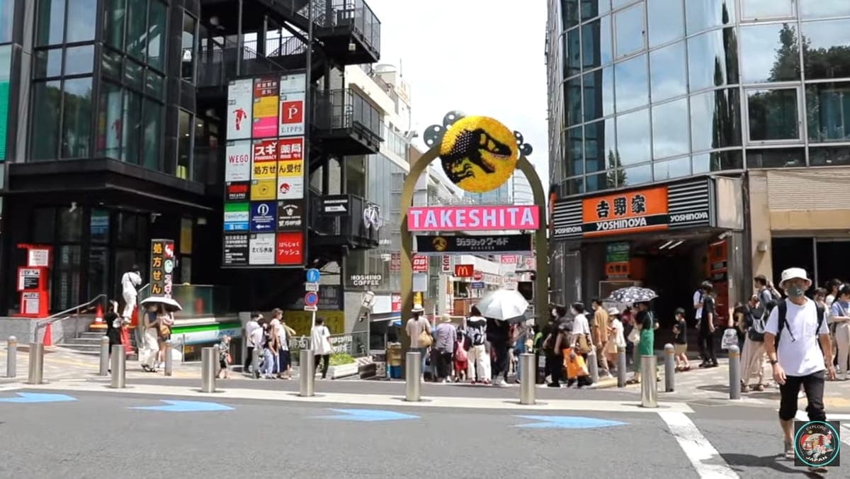 Vchod do ulice Takeshita v Harajuku s velkým obloukem s nápisem Takeshita, nápisy obchodů na skleněných budovách a nakupující přecházející silnici