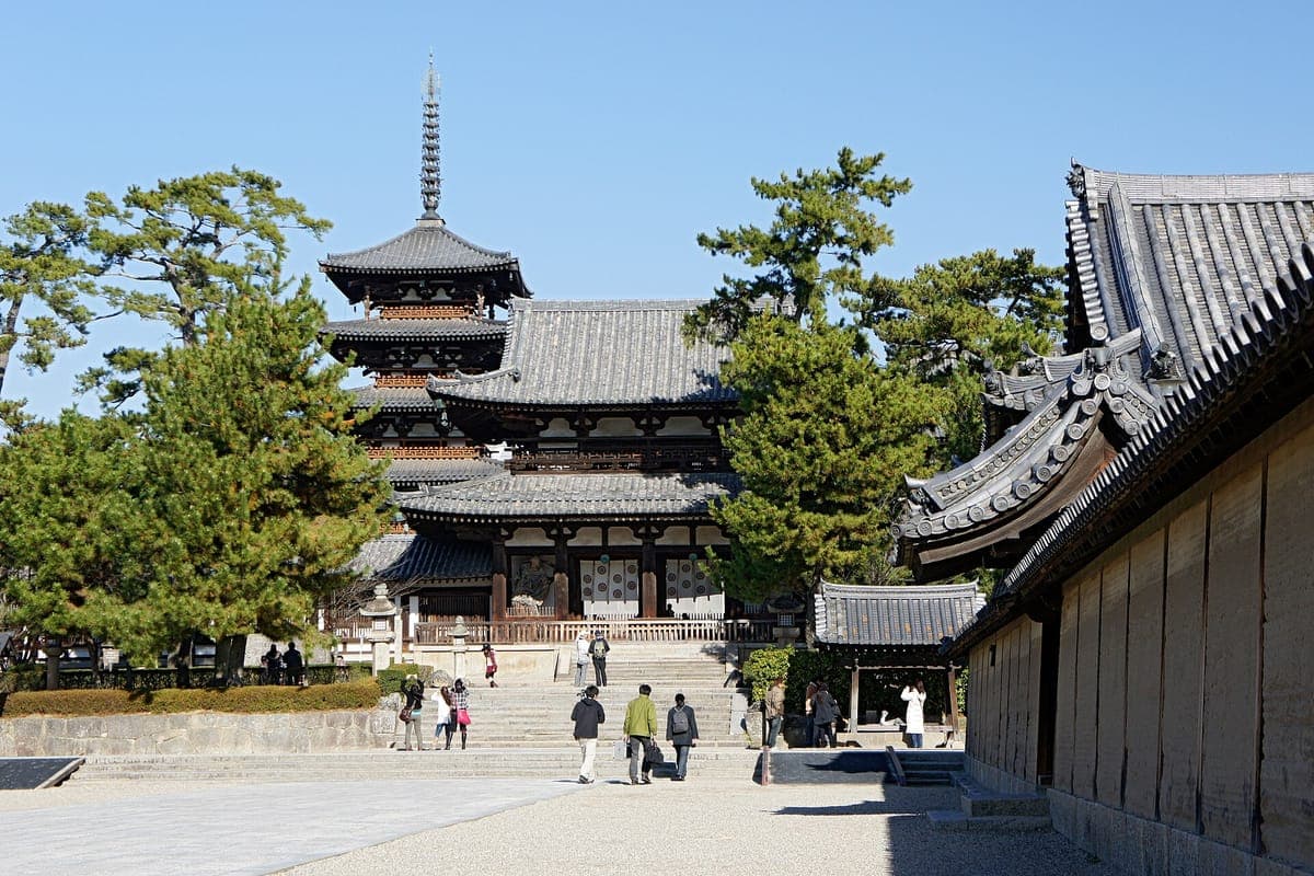 Un large chemin de pierre menant à la porte du milieu de Hōryū-ji, avec la pagode à cinq étages s'élevant derrière le cloître et les pins