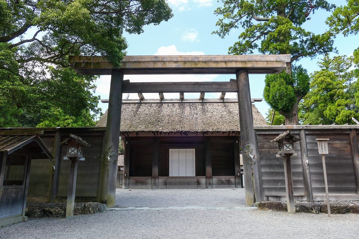 Un simple torii de madera sin pintar frente a una puerta con un grueso techo de paja y vallas de madera en Gekū, el Santuario Exterior de Ise Jingū