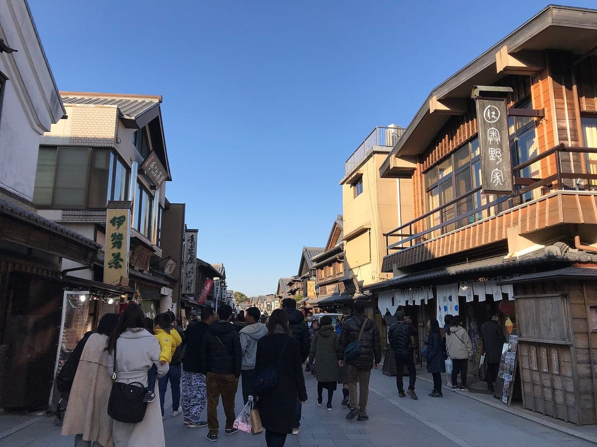 Los visitantes caminan por la calle Oharai-machi en Ise, pasando por tradicionales edificios comerciales de madera con techos de tejas y carteles colgantes bajo un cielo azul.