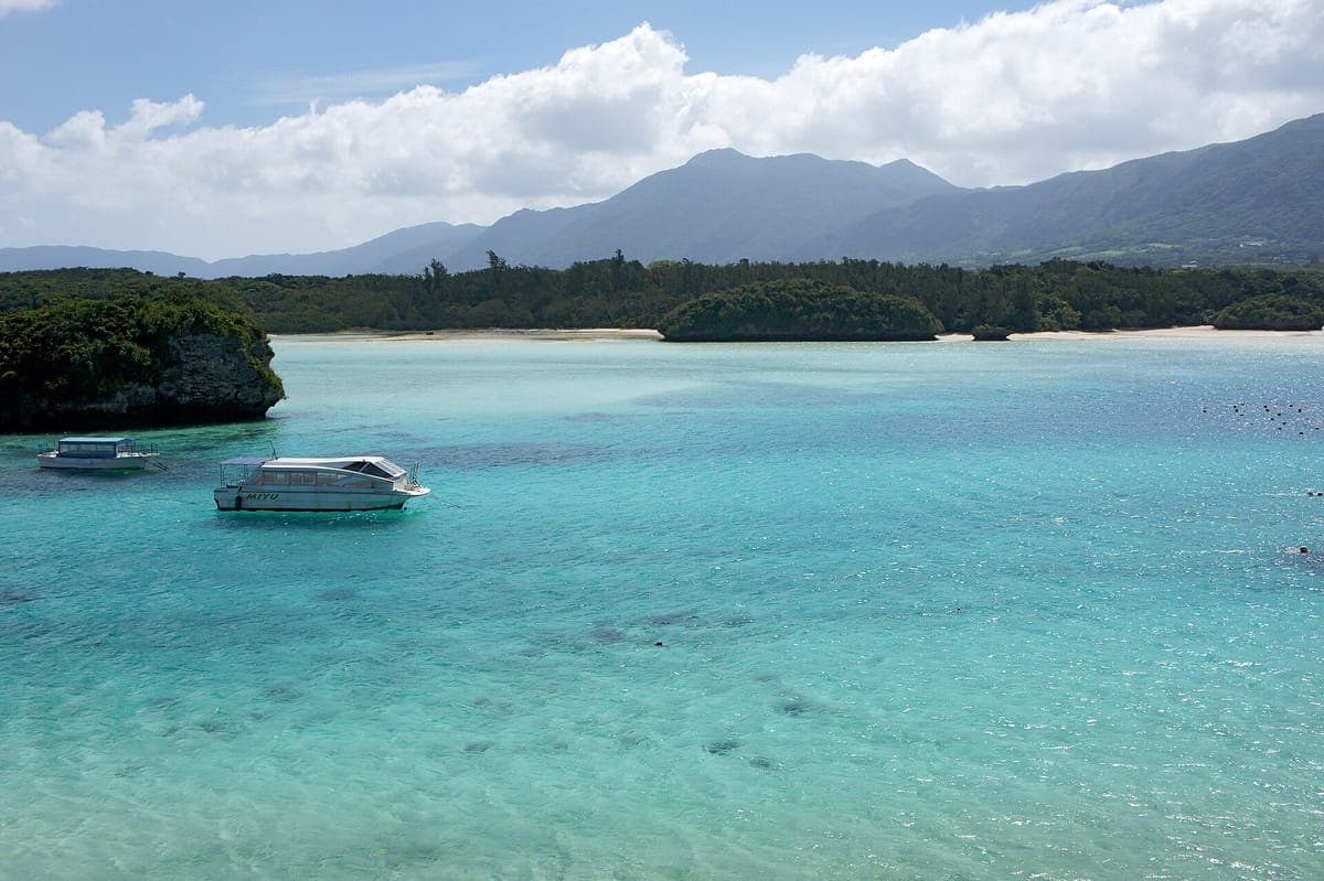 Das türkisfarbene Wasser der Kabira-Bucht auf der Insel Ishigaki mit kleinen Booten, die in der Nähe grüner, bewaldeter Inseln und dahinter liegender Berge festmachen