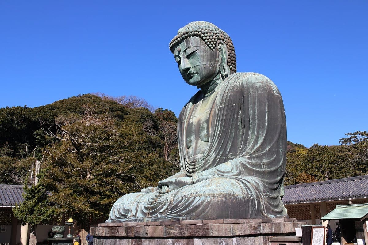 Bronzový Velký Buddha z Kamakury sedící na kamenné základně pod jasnou modrou oblohou se zelenými stromy a střechami chrámů za