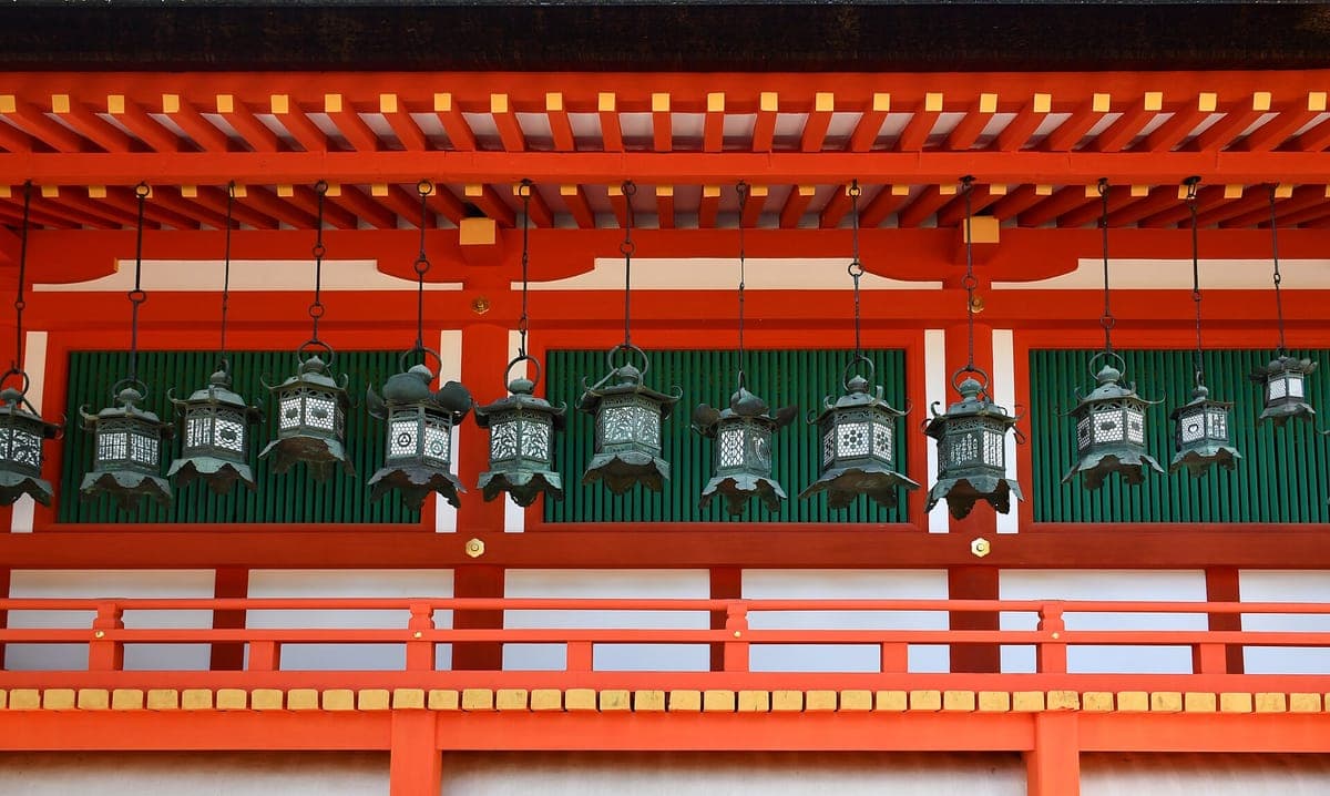 Kasuga Taisha အဆောက်အဦတစ်ခု၏ ဟင်္သပဒါးရောင် တံစက်မြိတ်အောက်တွင် ကြေးစိမ်းရောင်တွဲလောင်း မီးပုံးတန်းများ