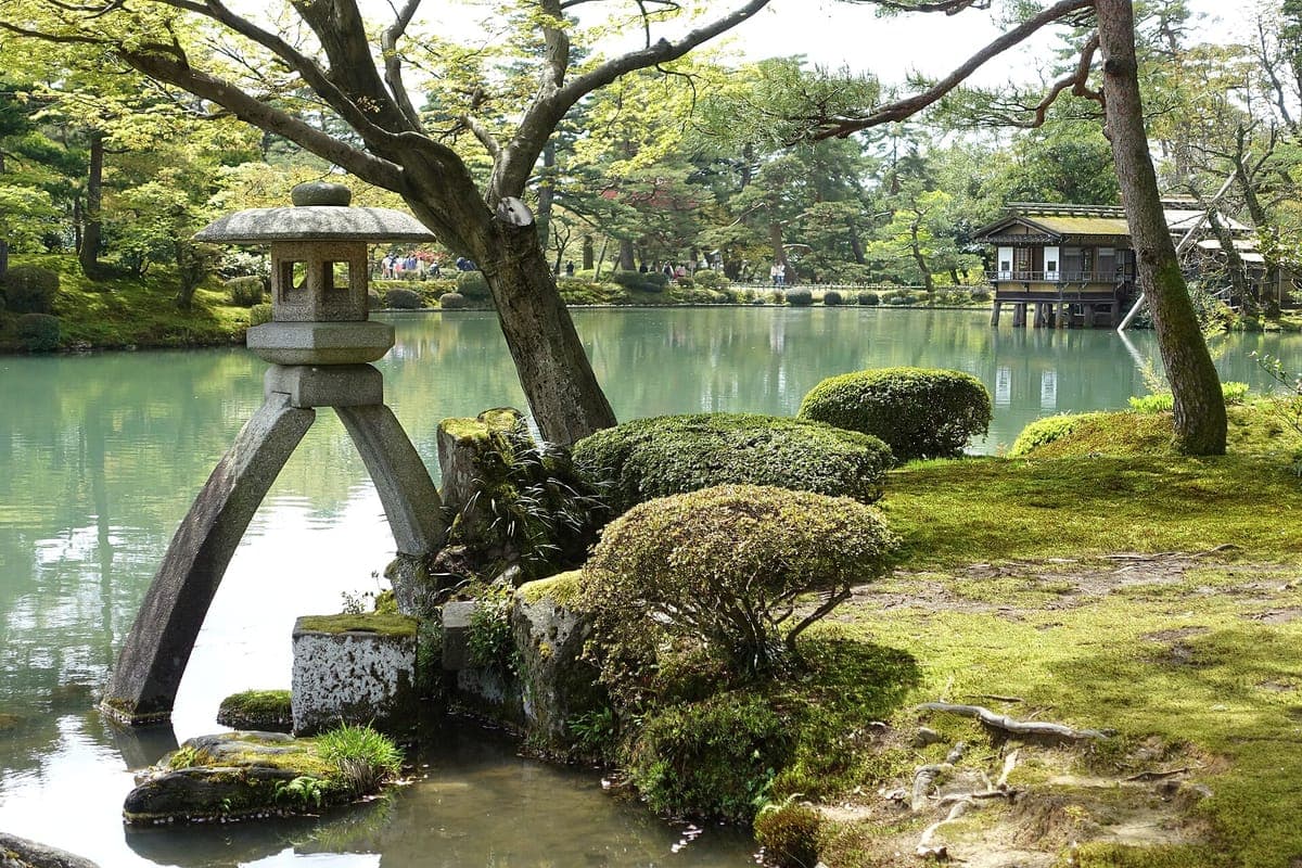 Ang dalawang paa na Kotoji stone lantern sa gilid ng Kasumiga-ike pond sa Kenroku-en, na may mga pinutol na palumpong, mga sanga ng pine at isang tea house sa kabila ng tubig