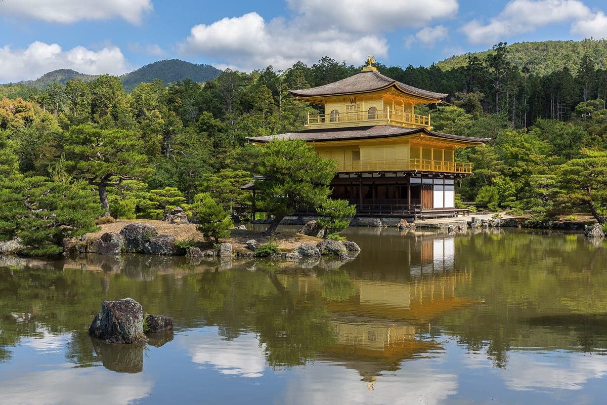 Ang gold-leafed na Kinkaku-ji pavilion sa gilid ng Kyōkochi pond, na masasalamin sa tubig sa ilalim ng asul na kalangitan na may puting ulap at mga burol sa likod.