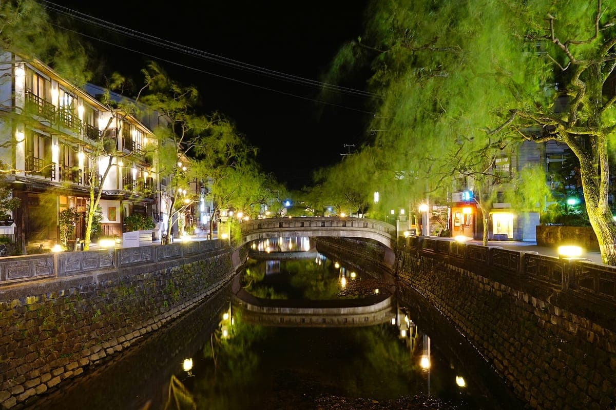 Kamenný kanál Kinosaki Onsen v noci, osvětlený pouličními lampami a okny hostince, se smutečními vrbami a klenutým kamenným mostem odrážejícím se ve vodě