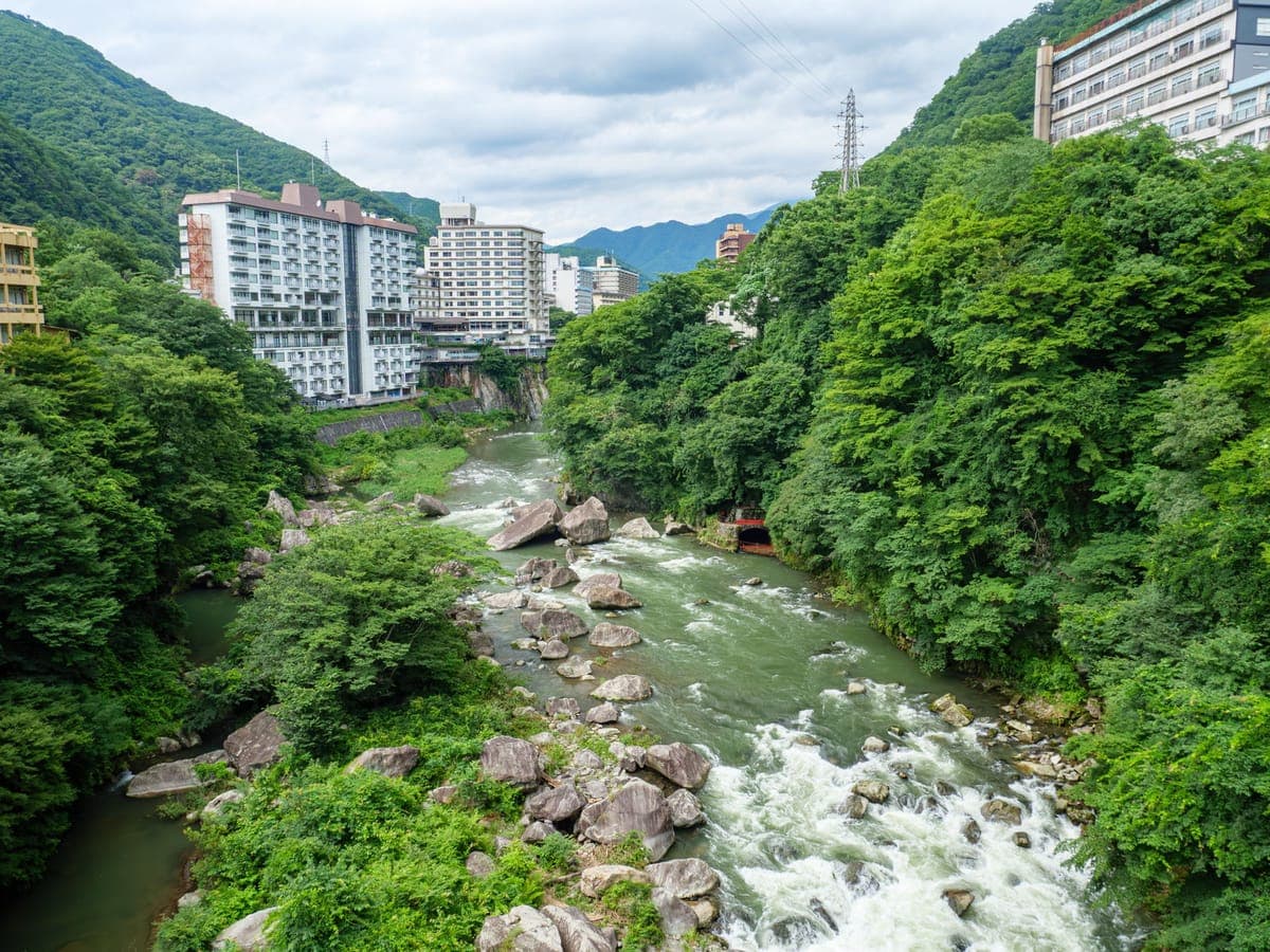 Řeka Kinugawa se řítí přes balvany zelenou zalesněnou roklí s velkými hotelovými budovami na vrcholcích útesů po obou stranách a horami za nimi