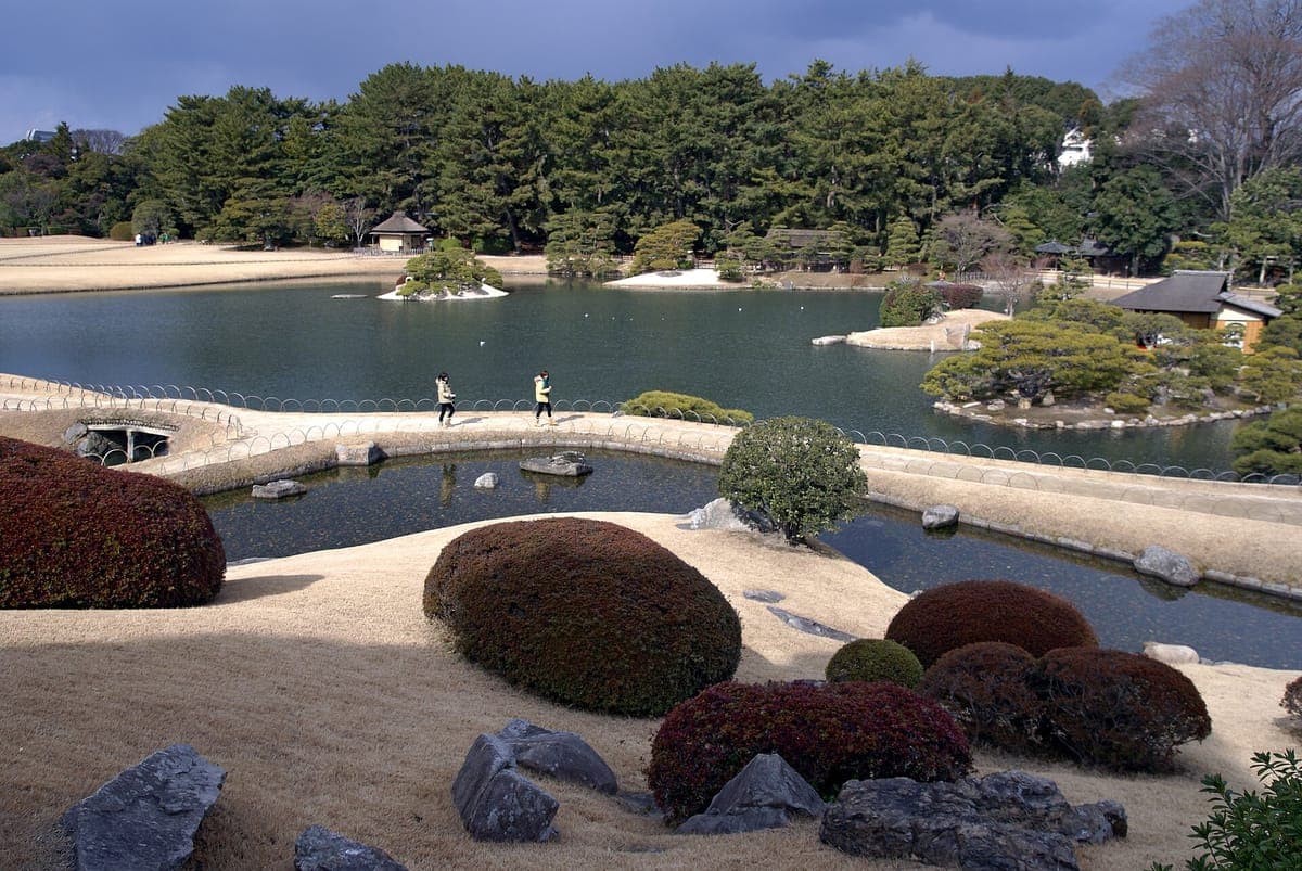 Kōraku-en ရှိ ကျွန်းများပါရှိသော ရေကန်ကြီးတစ်ခု၏အထက် သဲသောင်ပြင်ပေါ်တွင် အဝိုင်းပတ်ထားသော ချုံဖုတ်များနှင့် ကျောက်တုံးများ၊ ထင်းရှူးပင်များနှင့် ကမ်းစပ်ရှိ မဏ္ဍပ်ငယ်လေးများပါရှိသည်။