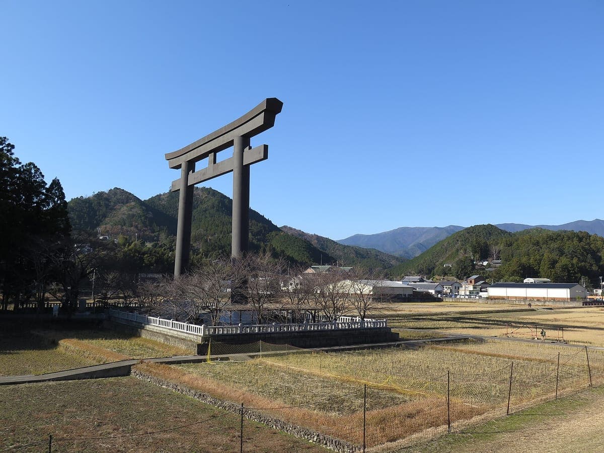 L'énorme porte torii sombre à Ōyunohara se dressant au-dessus de champs plats avec des montagnes boisées et un ciel bleu derrière