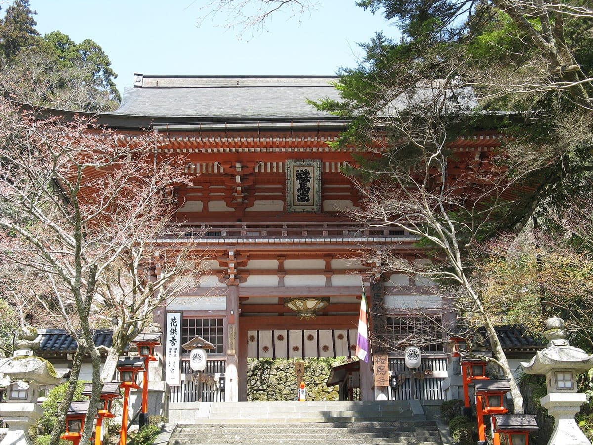 O portão vermelho de dois andares Niōmon de Kurama-dera no topo de degraus de pedra, com lanternas vermelhas e árvores nuas em ambos os lados