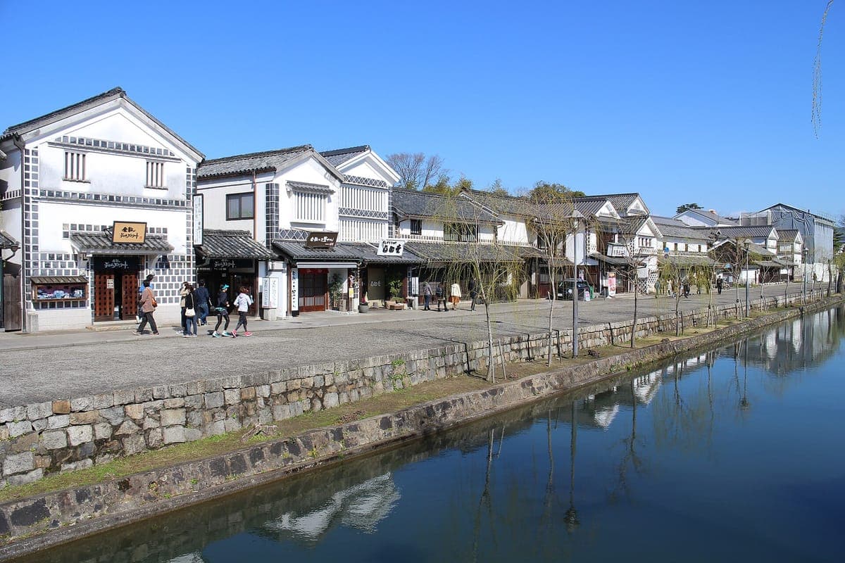 Le canal Kurashiki avec une digue en pierre et une eau toujours bleue, bordé d'entrepôts aux murs blancs avec des toits de tuiles noires et des saules nus sous un ciel clair