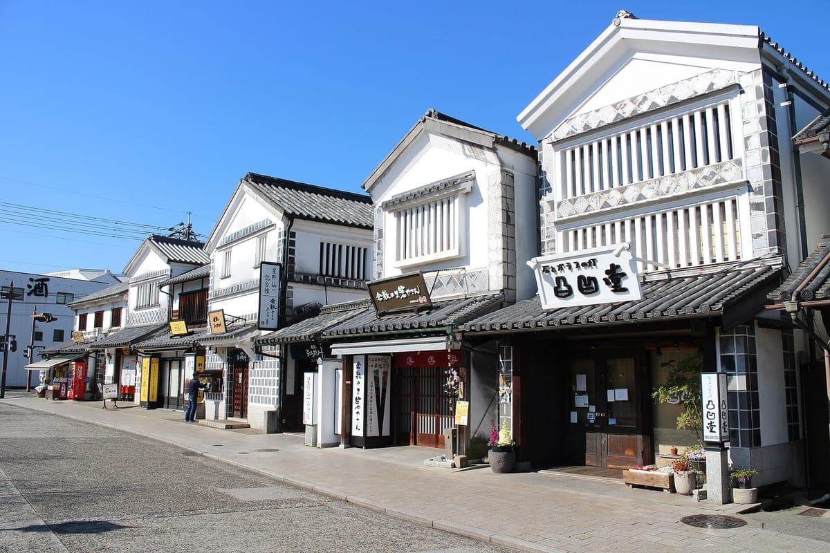 Une rue du quartier Kurashiki Bikan bordée d'entrepôts en plâtre blanc avec des toits de tuiles noires, des fenêtres en treillis et des enseignes de magasins, sous un ciel bleu clair