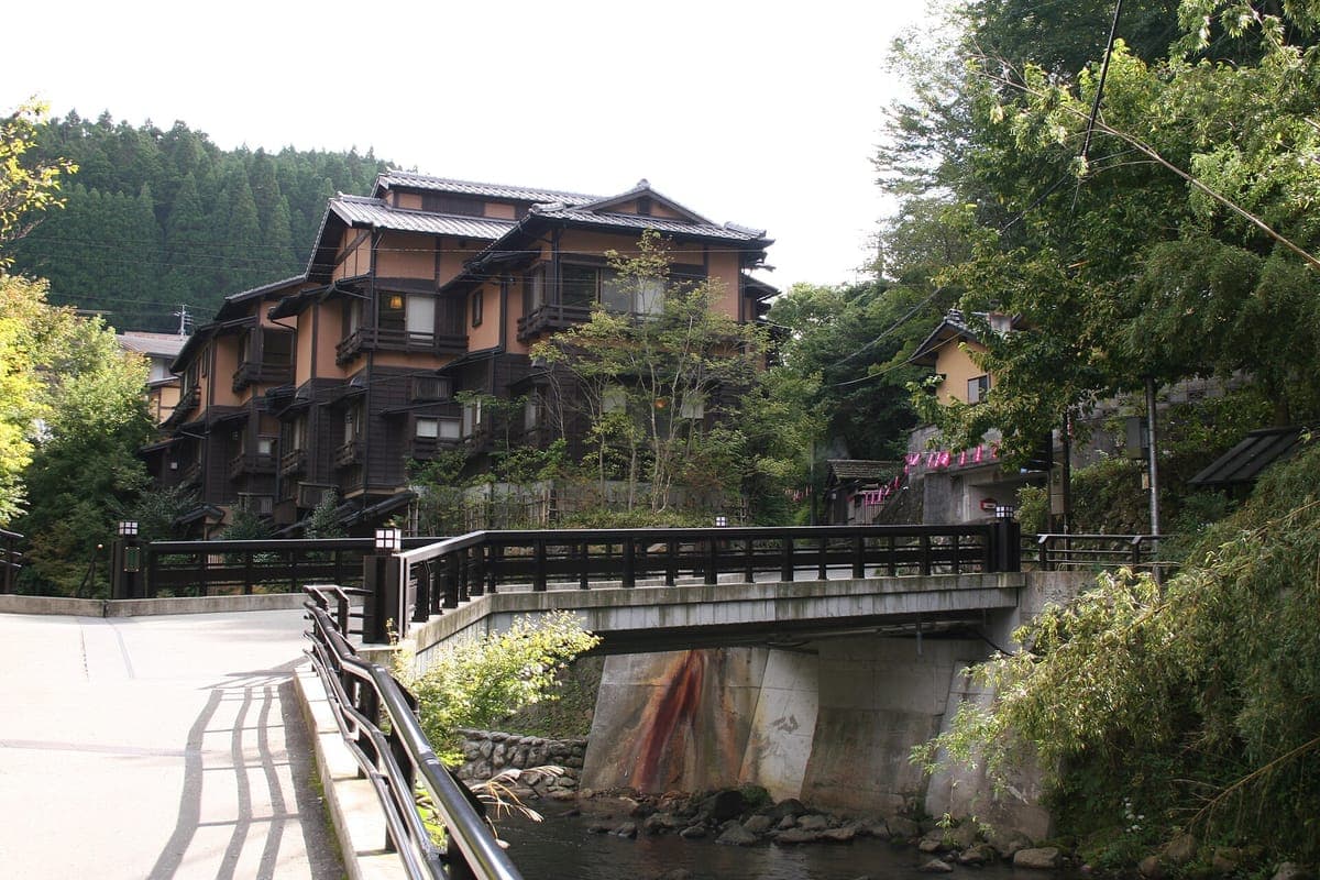 Isang tatlong palapag na ryokan na gawa sa kahoy na may madilim na mga balkonaheng gawa sa kahoy sa tabi ng isang maliit na tulay sa ibabaw ng batis sa berdeng lambak ng Kurokawa Onsen