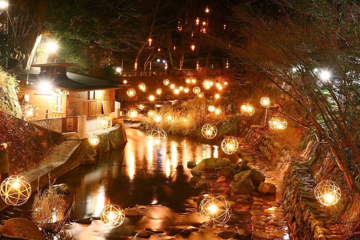 Mga bilog na parol na kumikinang na orange sa tabi ng batis sa gabi sa Kurokawa Onsen, na sinasalamin sa tubig sa ilalim ng nakasinding kahoy na gusali