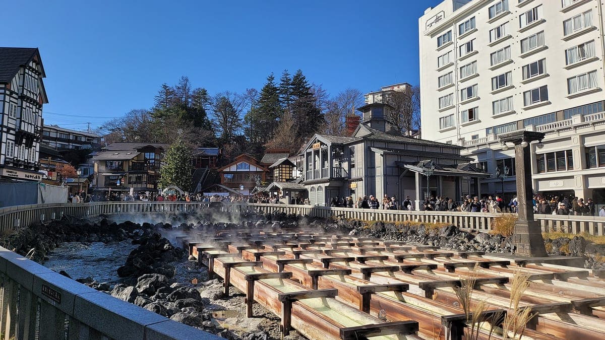 Ang Yubatake sa Kusatsu Onsen: mga hilera ng mga channel na gawa sa kahoy na nagdadala ng umuusok na mainit na bukal na tubig sa ibabaw ng mga bato, na napapalibutan ng isang batong rehas, mga hotel at mga gusaling gawa sa kahoy