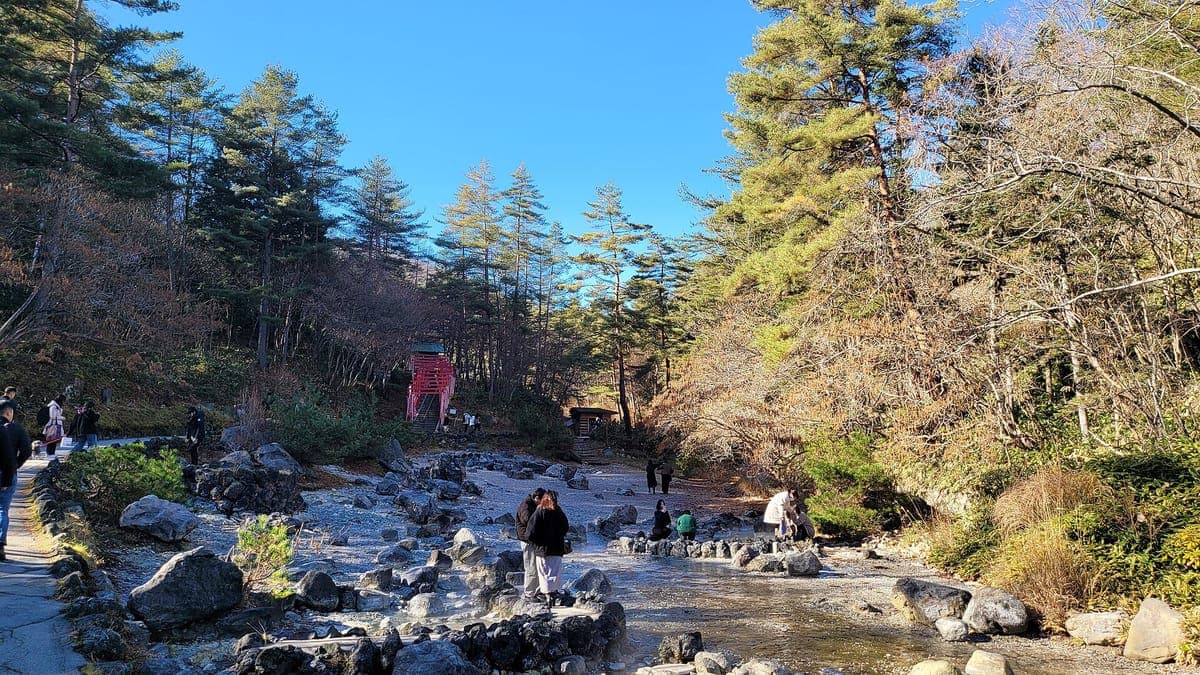 Isang mabatong stream bed sa Sainokawara Park sa Kusatsu na may umuusok na mainit na tubig, mga pine at taglagas na puno sa mga pampang at isang maliit na pulang shrine sa di kalayuan