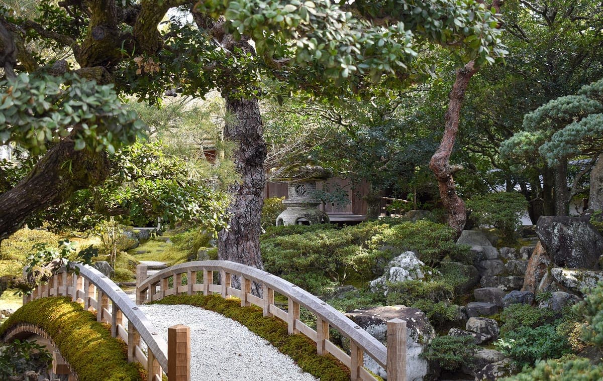 Il giardino di un palazzo con un ponte di legno ad arco dolce, ghiaia rastrellata, lanterne di pietra e pini potati con cura
