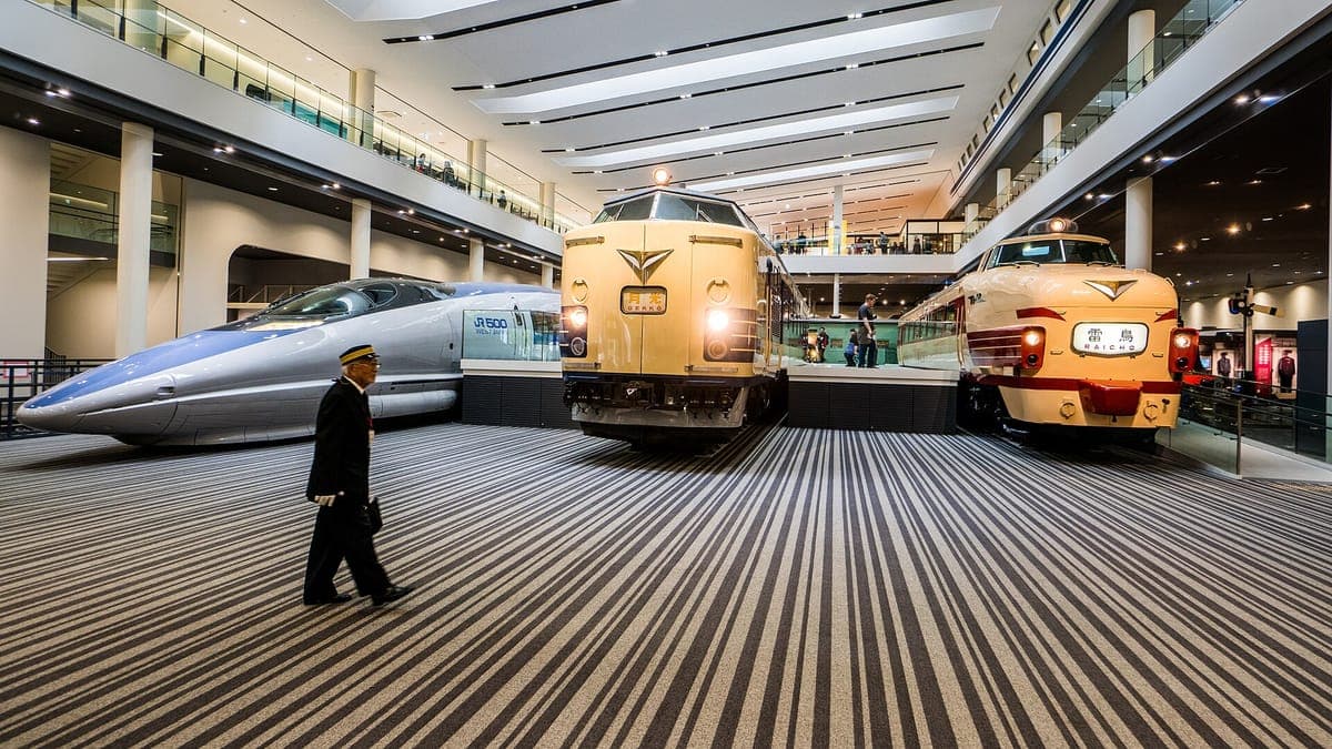 Dentro de la sala principal del Museo del Ferrocarril de Kioto, una nariz puntiaguda blanca y azul shinkansen junto a dos trenes expresos color crema sobre un suelo rayado, con un hombre caminando