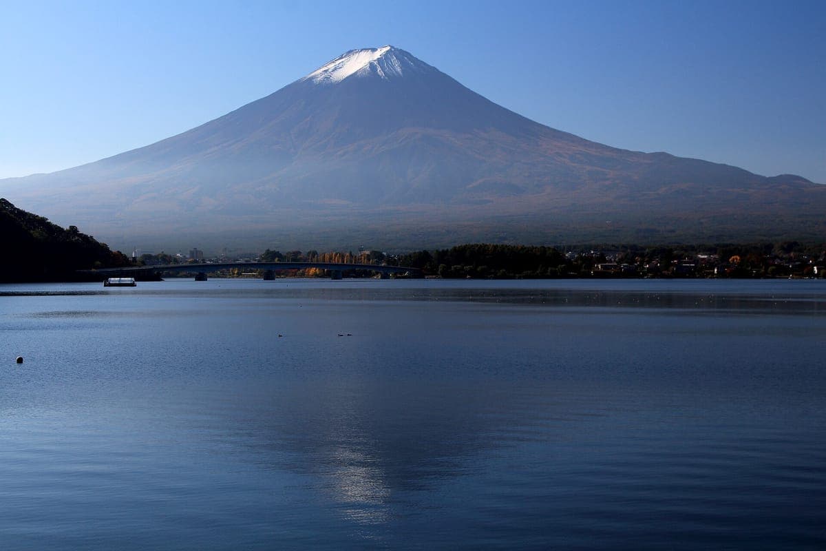 Góra Fuji z lekką czapą śniegu wznoszącą się nad spokojną błękitną wodą jeziora Kawaguchi, z słabym odbiciem i zalesionym brzegiem