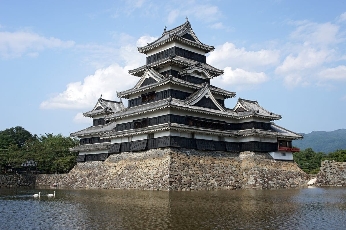 Der schwarz-weiße fünfstufige Bergfried der Burg Matsumoto auf seinem Steinsockel, der sich im Wassergraben unter einem blauen Himmel mit weißen Wolken spiegelt