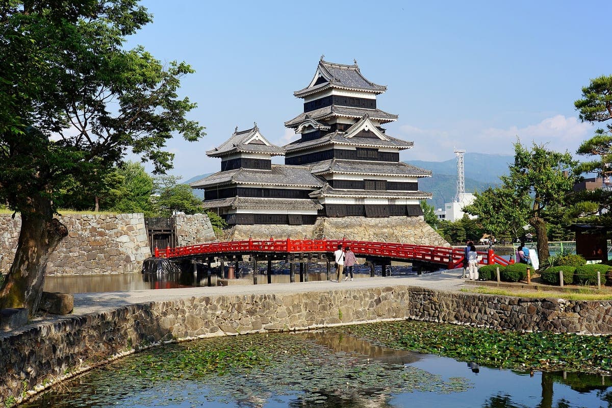Eine rot gestrichene Holzbrücke überquert den grünen Wassergraben zum schwarzen Bergfried der Burg Matsumoto, eingerahmt von Bäumen und einer Steinmauer