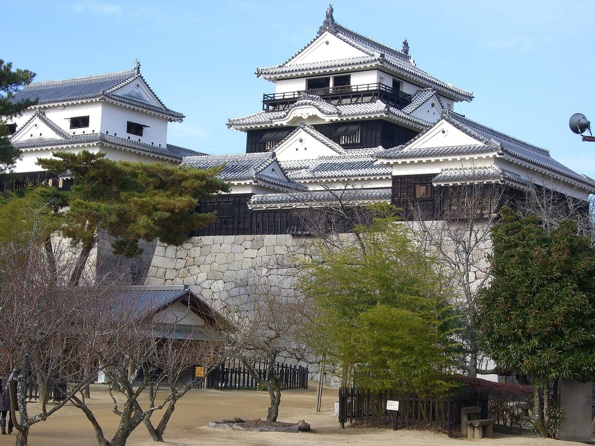 La torre principal del Castillo de Matsuyama, de paredes blancas y madera negra, con techos en capas, conectados a torres más pequeñas sobre altos muros de piedra.