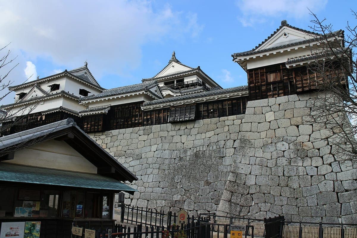 Altos muros curvos de piedra del castillo de Matsuyama coronados por torreones blancos y la torre del homenaje, con un pequeño edificio de madera en primer plano