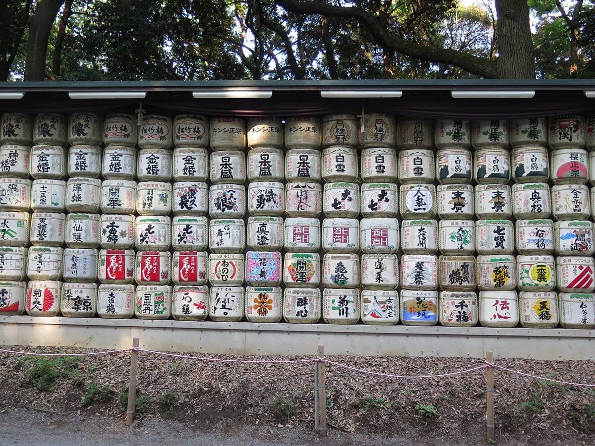 Un long mur de fûts de saké décorés empilés en rangées sous un toit à côté d'un chemin bordé d'arbres à Meiji Jingū