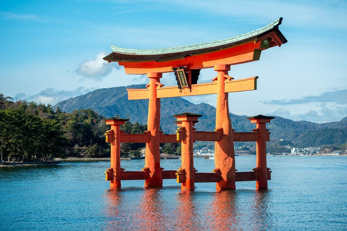 Torii besar kuil Itsukushima yang berwarna merah jingga berdiri di dalam air biru yang tenang dengan pergunungan berhutan di belakang