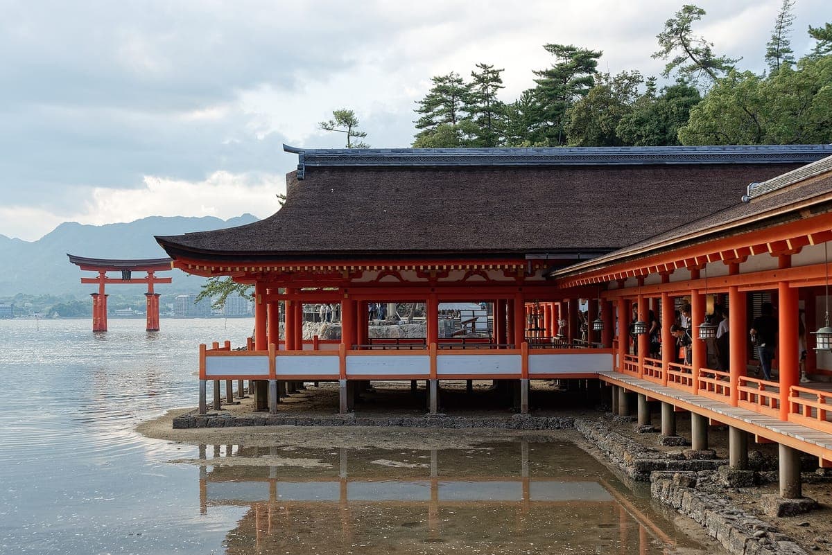 Koridor vermilion dan bumbung gelap Kuil Itsukushima berdiri di atas timbunan pasir basah dan air cetek, dengan torii besar di teluk di belakang