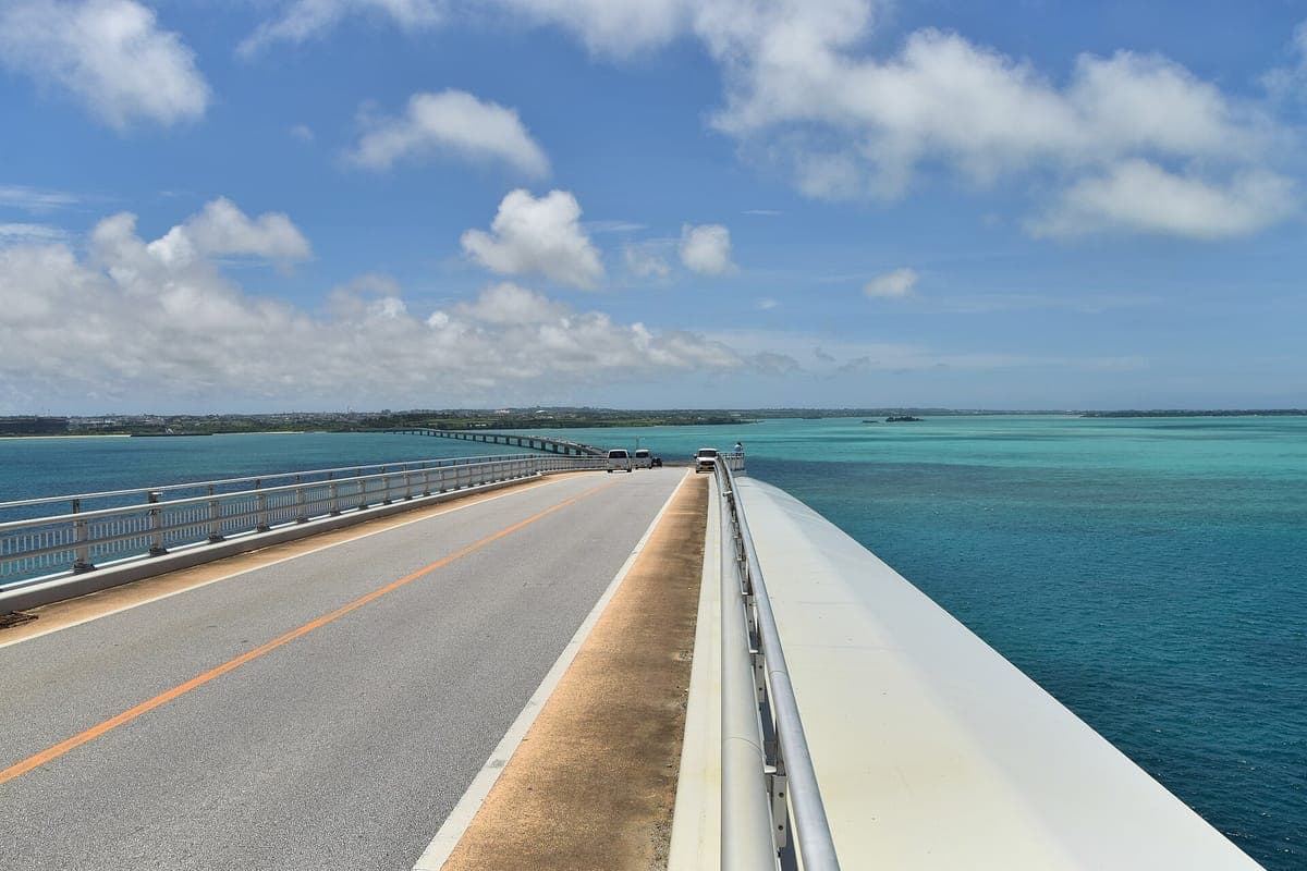 Die lange Irabu-Brücke erstreckt sich über türkisfarbenes, flaches Wasser zu einer niedrigen grünen Insel unter einem blauen Himmel mit weißen Wolken