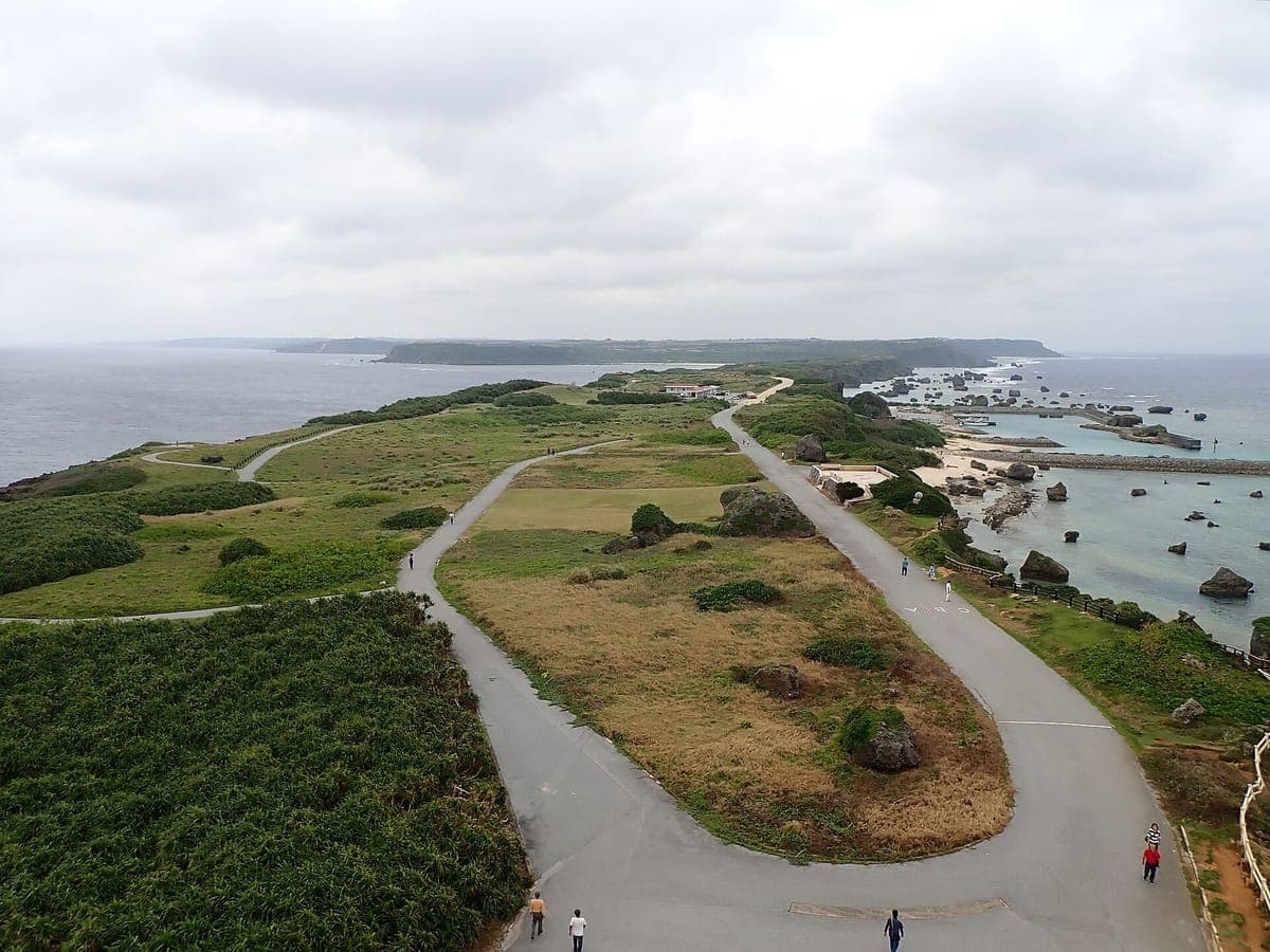 Das lange, schmale Kap von Higashi-Hennazaki von oben gesehen, mit einer kurvenreichen Straße und Pfaden über grasbewachsenes Land zwischen dem Meer auf beiden Seiten