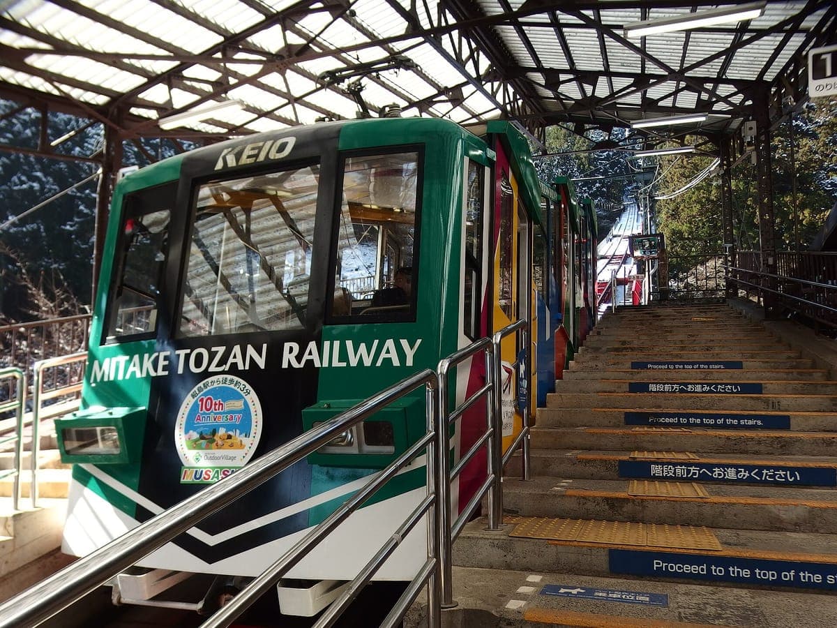 Eine grün-weiße Seilbahn der Mitake Tozan Railway wartet an einer überdachten Station neben einem abgestuften Bahnsteig mit einer aufgemalten Führungslinie