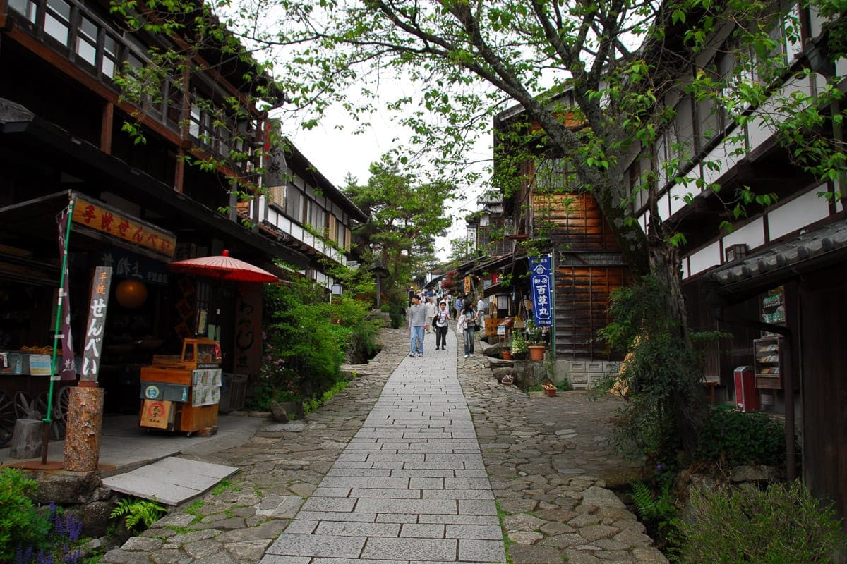 La pendiente empedrada de Magome-juku trepa entre antiguas tiendas de madera y casas con plantas, faroles y una sombrilla roja a lo largo del camino.