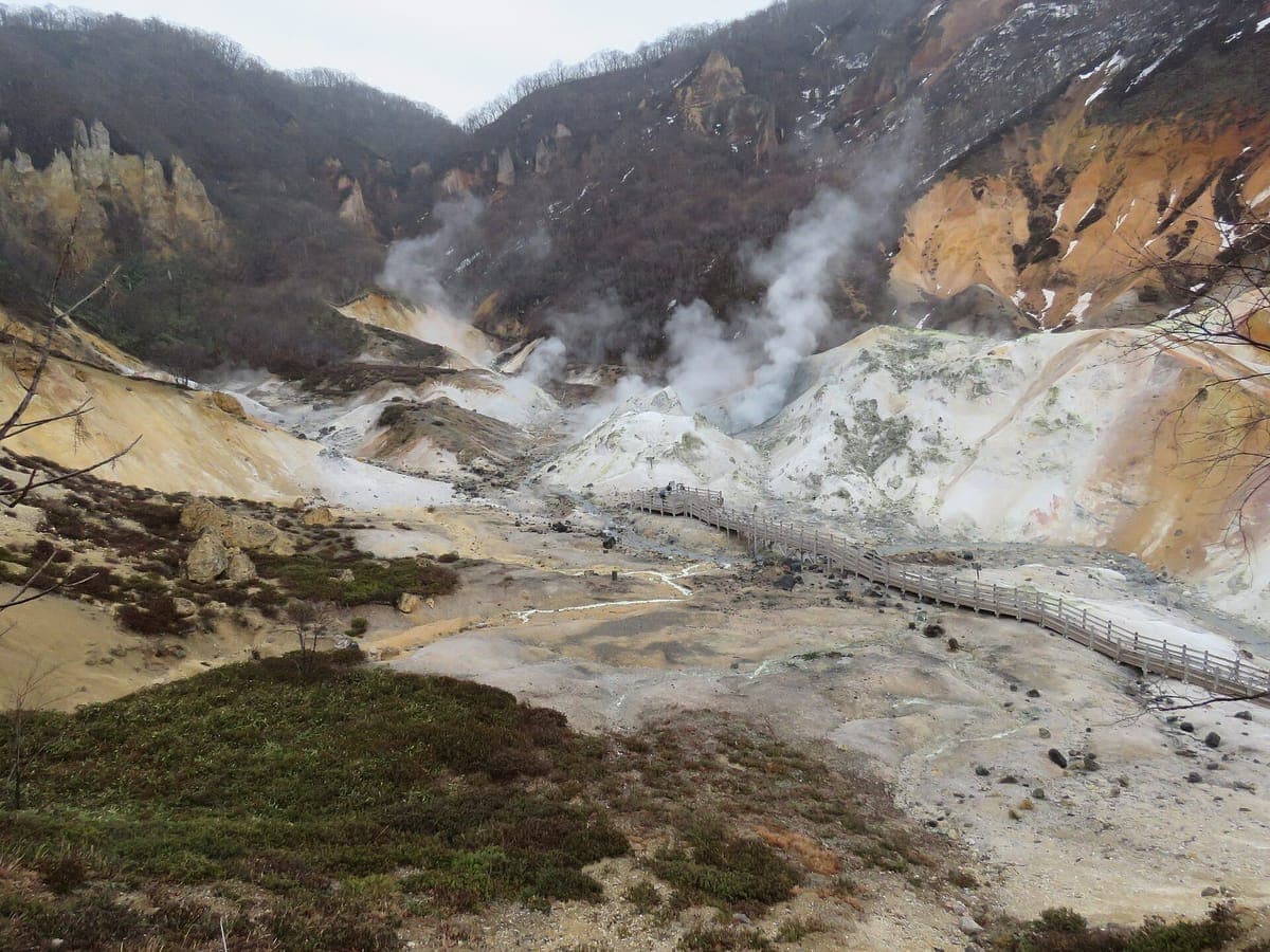 Jigokudani in Noboribetsu, einem Tal aus blassgelbem und orangefarbenem Vulkangestein, durch das sich Dampf aus Öffnungen aufsteigt und durch das sich ein Holzsteg schlängelt