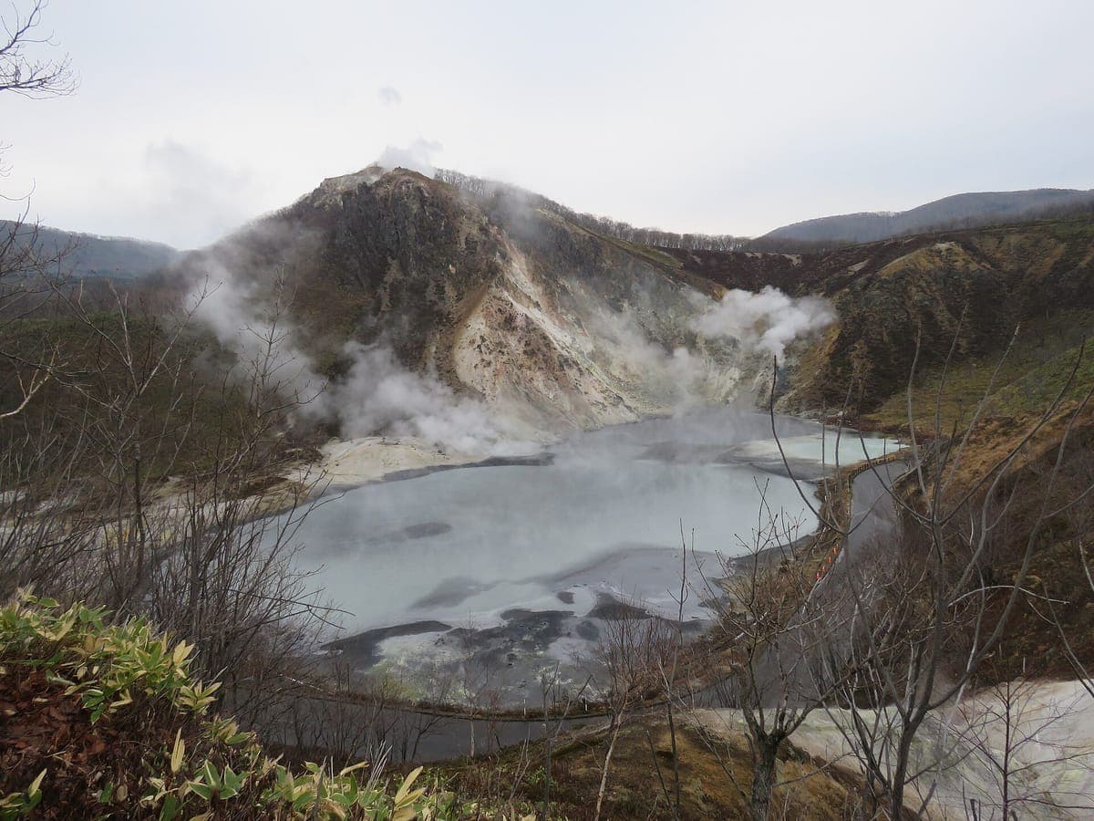 Ōyunuma Teich in Noboribetsu, ein großer grauer Schwefelteich, an dessen gegenüberliegendem Ufer unterhalb eines felsigen Vulkanhügels Dampf aufsteigt, umgeben von kahlen Bäumen