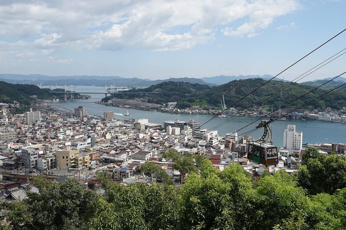 Pohled z kopce Senkō-ji přes střechy Onomichi na kanál a ostrov Mukaishima, s lanovkou na kabelech v popředí