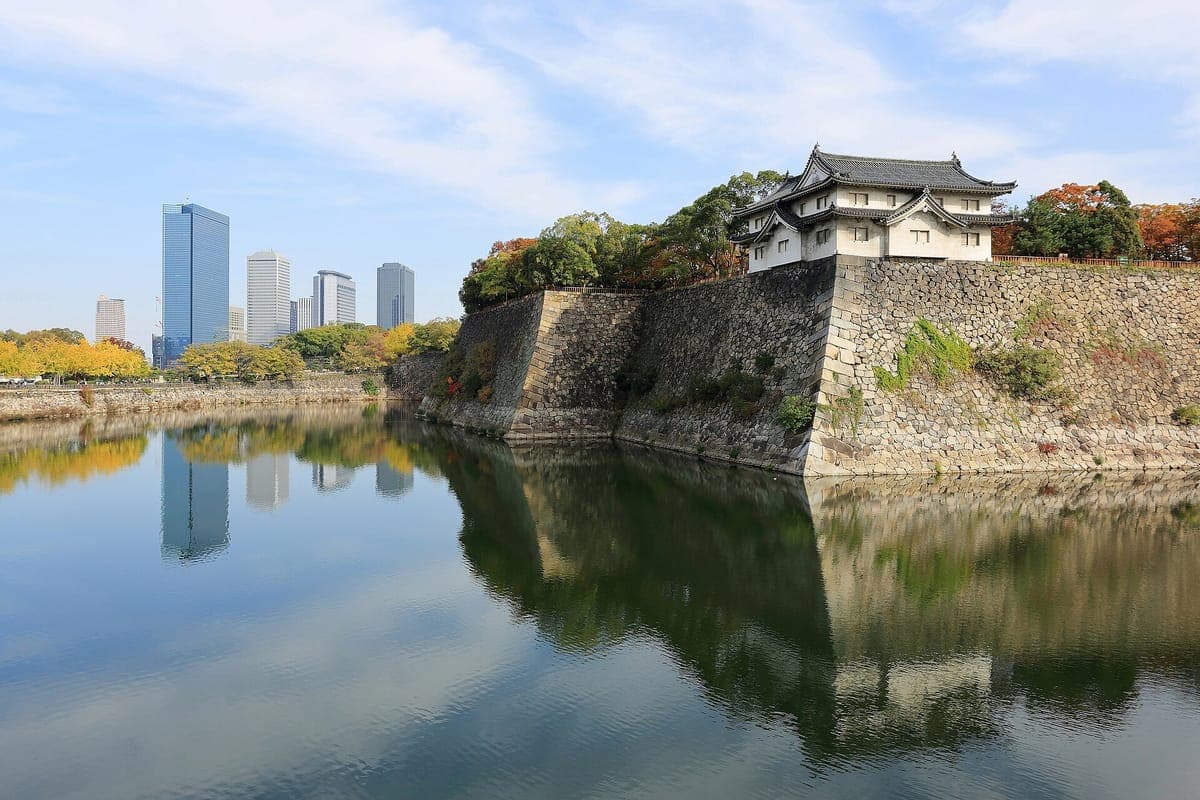 Un alto muro de piedra curvado y una torre blanca del Castillo de Osaka se reflejan en las tranquilas aguas del foso exterior, con torres de oficinas al fondo